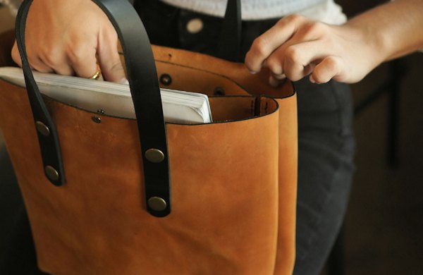 Mãos guardando um caderno em uma bolsa tote de couro caramelo com alças pretas